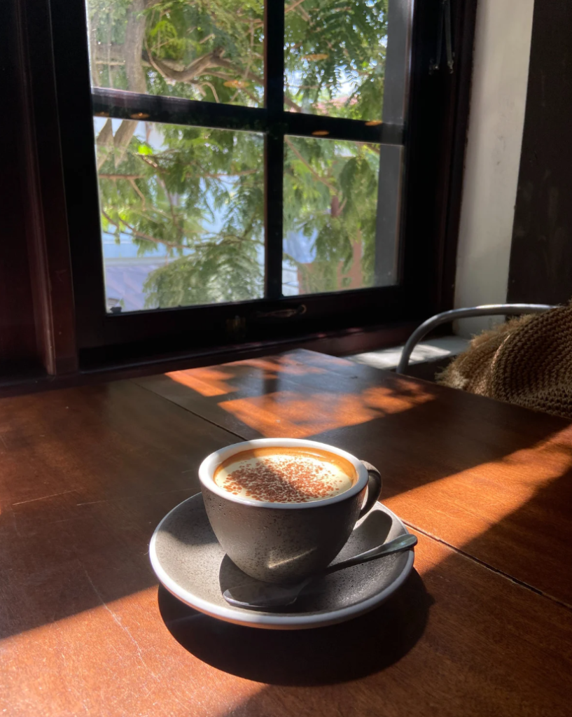 Frau sitzt morgens am Küchentisch mit einer Tasse Kaffee im Sonnenlicht vor einem Fenster